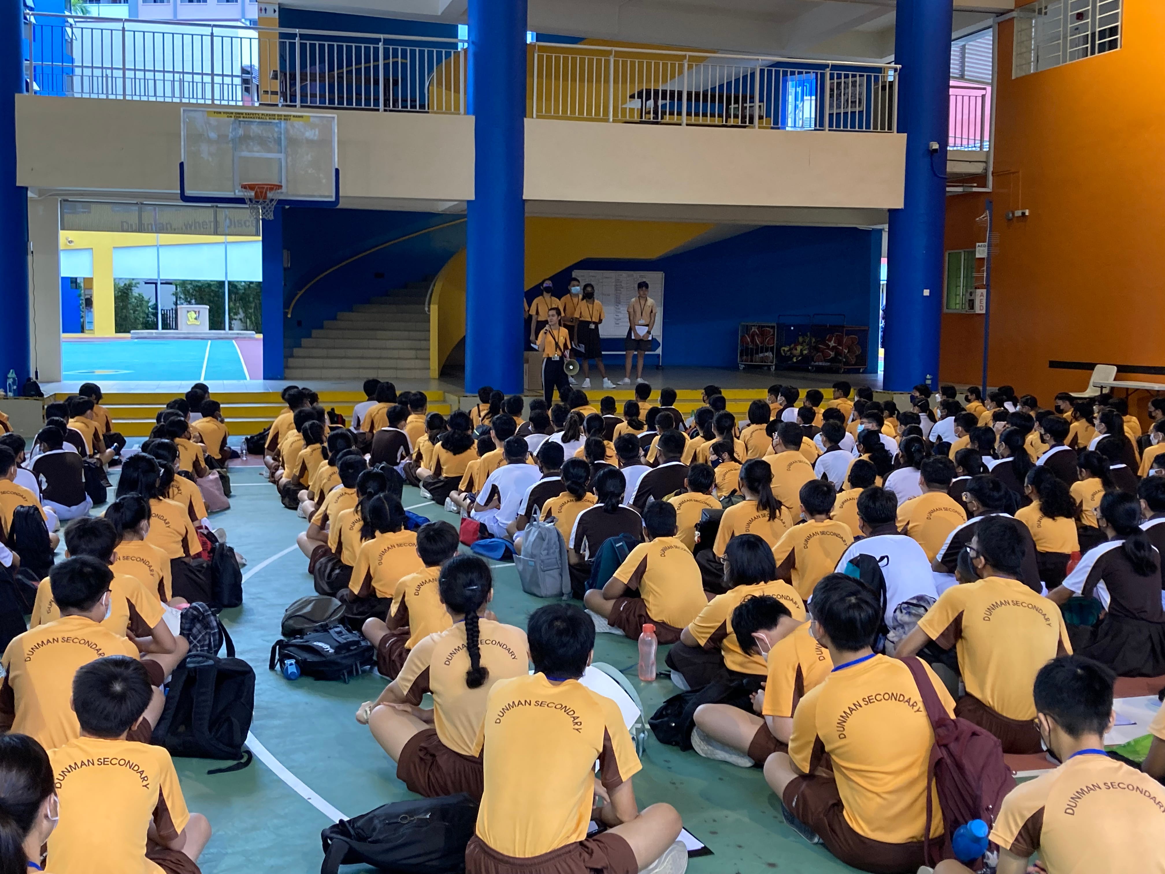 Students in yellow Dunman Secondary shirts sit on a gym floor facing speakers at the front of the hall.